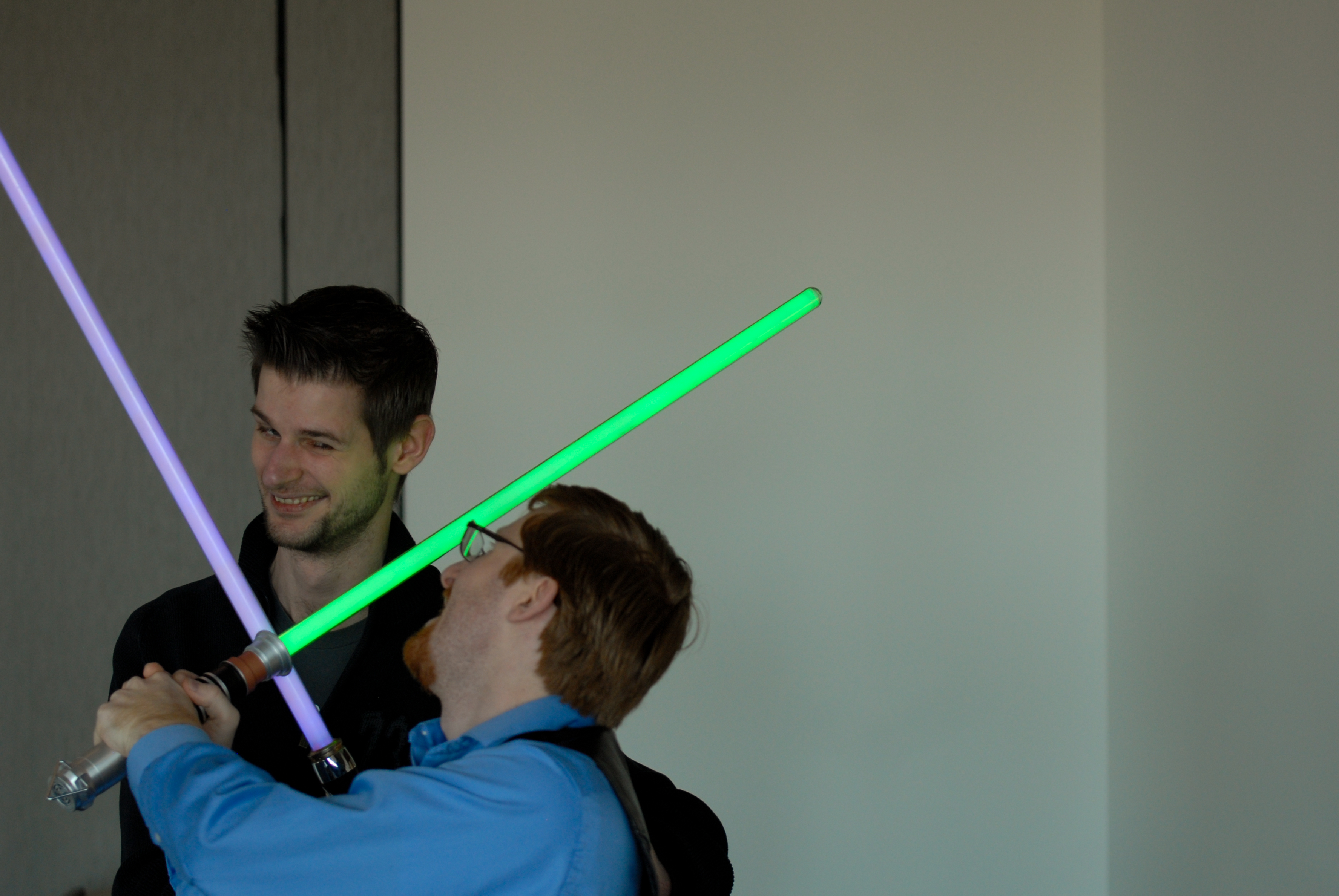 Two people smiling while playfully dueling with toy lightsabers, one green and one purple.